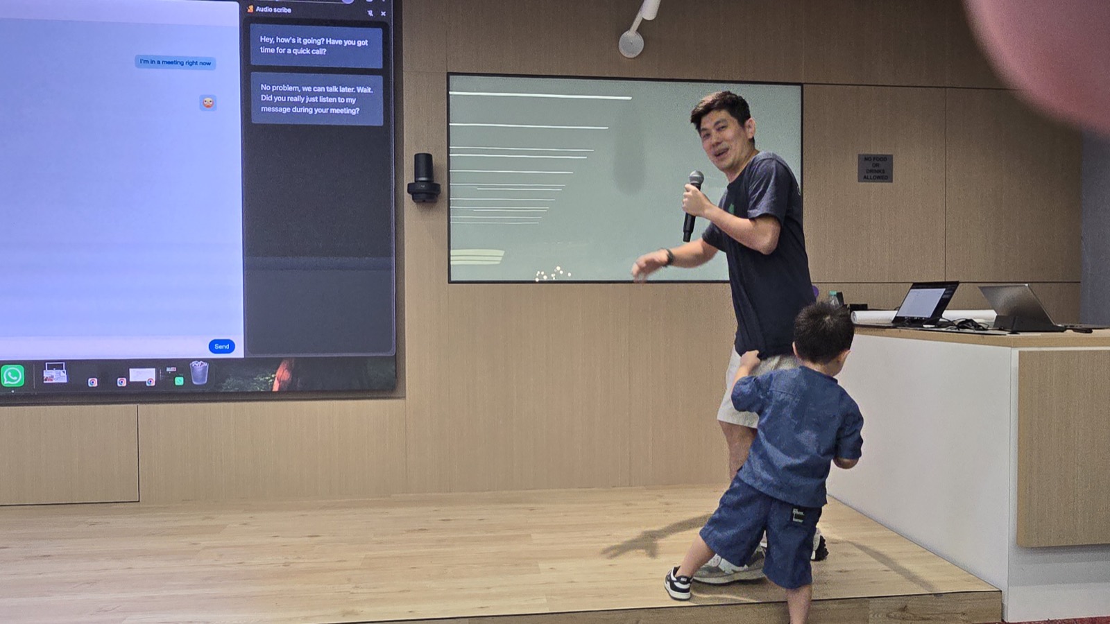 Vin Lim on stage at GDG Meetup #12 in Kuala Lumpur beside a screen showing the Audio Scribe side panel transcribing chat messages, with his young son on stage beside him
