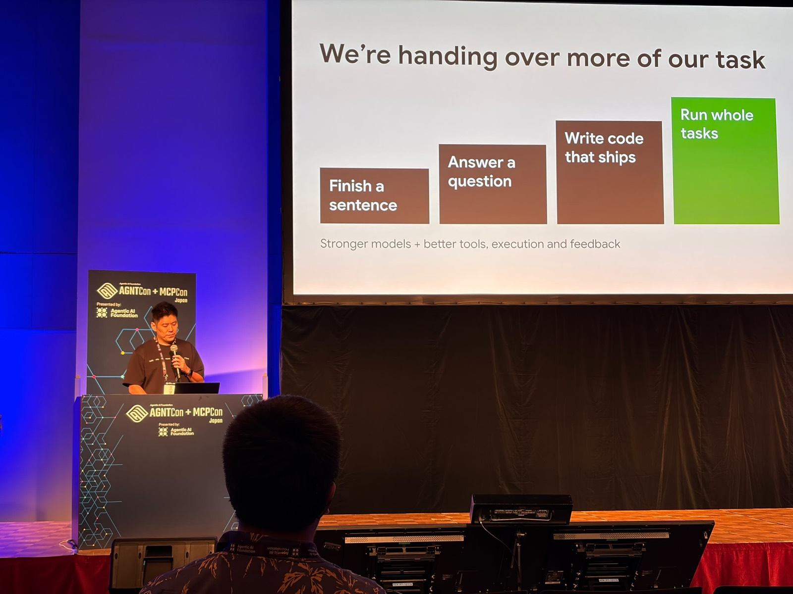 Vin Lim speaking at the AGNTCon + MCPCon Japan lectern in Tokyo, beside a slide titled We're handing over more of our task